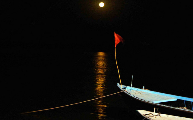 Image: A red flag on a boat in the water at night with the moon in the background
