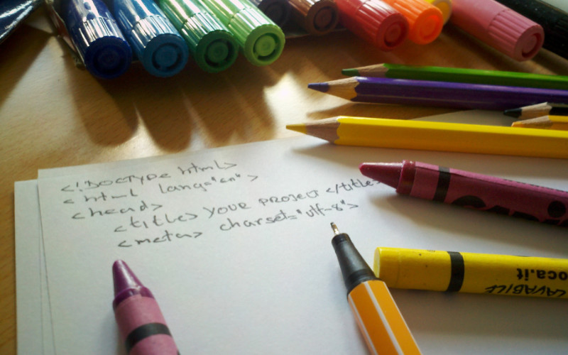 Image: Hand-written HTML code on paper surrounded by crayons, pens, and drawing implements.