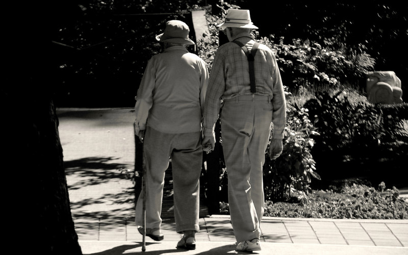 Image: Two elderly people walking away from the camera, holding hands.