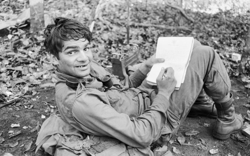 Image: A soldier lying down and writing on a notepad in the Vietnam war, black and white, surrounded by foliage.