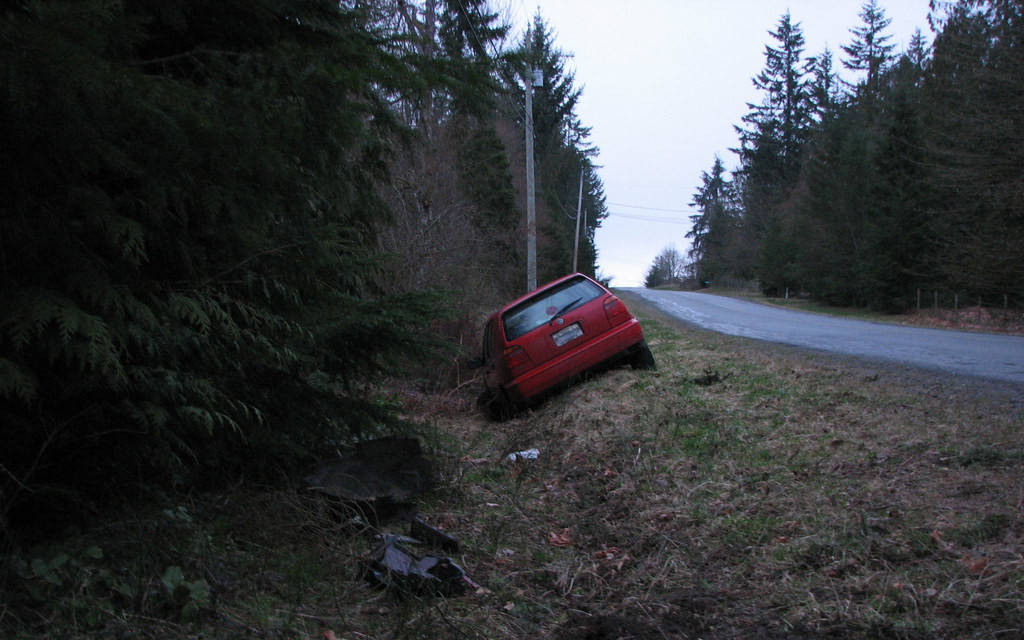 Image: Photo of a red car that has driven into a ditch on a road going through a pine forest