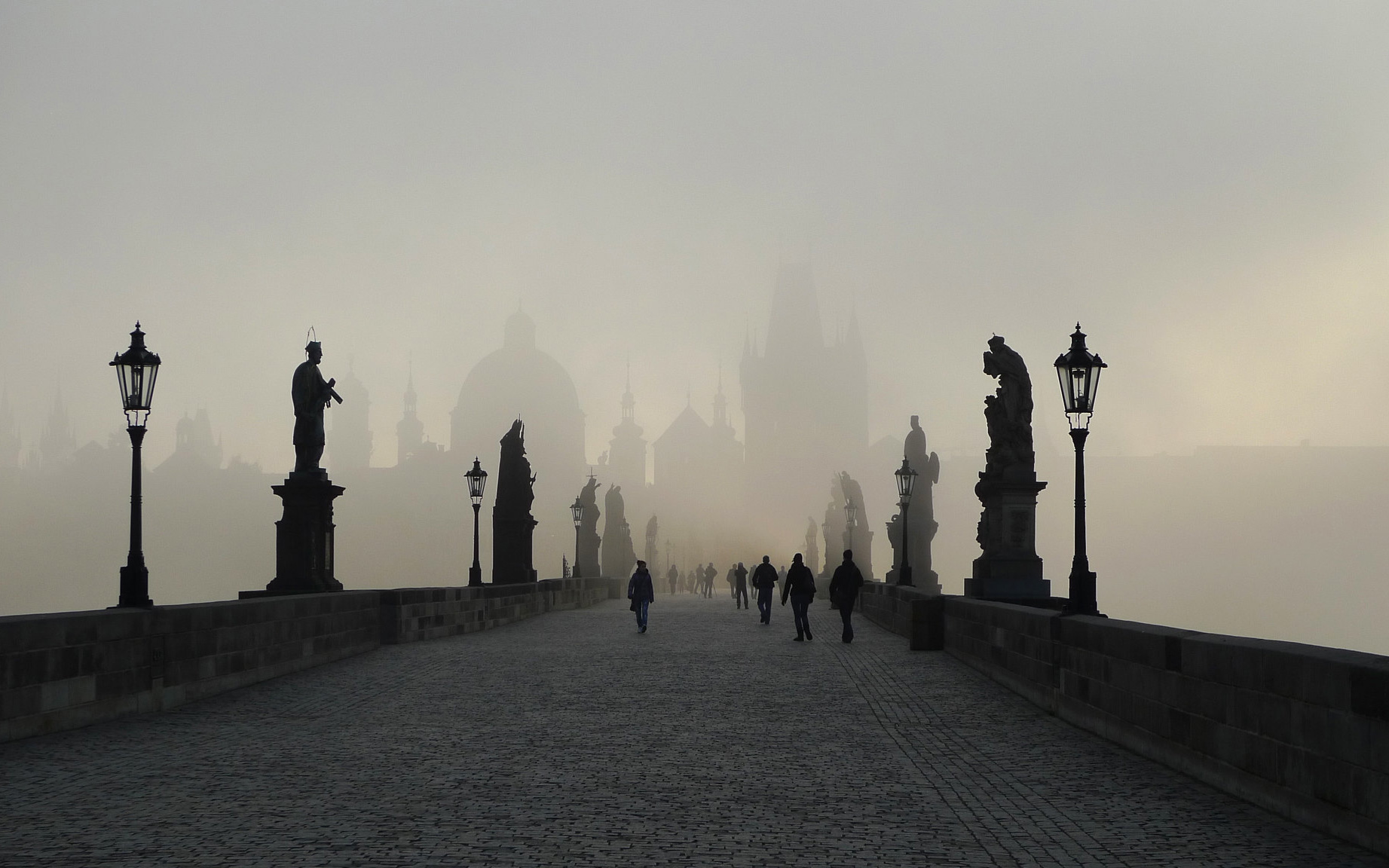 Image: Photo of a bridge on a foggy dawn. Silouhettes walk over the stone path under the water of statues. Titled, Prague: Charles Bridge in the Mist (Explored)