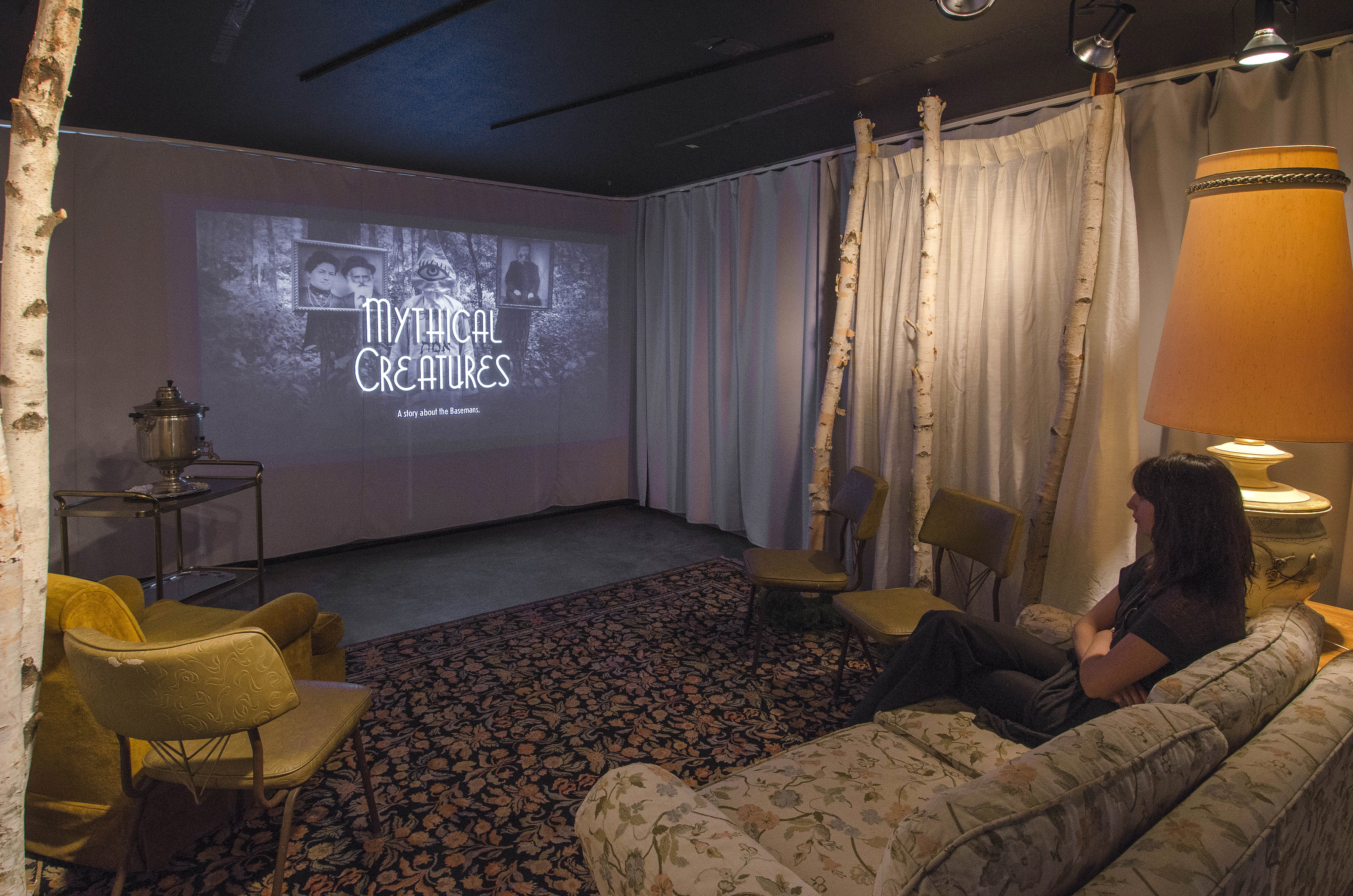Image: A woman sits on a couch watching a film by projector called "Mythical Creatures".
