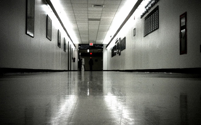 Image: A dark hallway in black and white with the exit sign still in red.